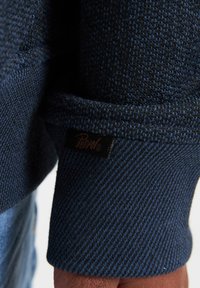 Felpa blu navy con tessuto testurizzato, polsini a coste e un'etichetta nera con la scritta "Perrico" sulla manica.