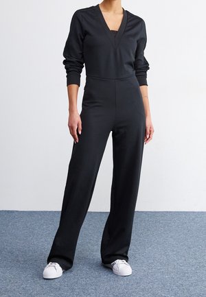 Woman wearing a black long-sleeve jumpsuit with a V-neck and white sneakers, standing on a gray carpet against a white background.