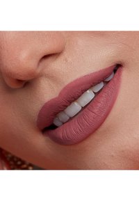 Matt läppstift i mjuk mauv-ton på fylliga läppar, som framhäver den släta texturen och vita tänder. Närbild på munnen med jämn färgfördelning.