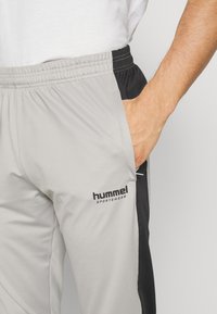 Grå och svarta sportbyxor med elastisk midja, sidofickor och "hummel SPORTWEAR"-logotyp på låret. Slät textur.