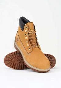 Stivaletti alla caviglia in suede beige con dettagli in pelle nera, occhielli dorati e lacci coordinati. Suola in gomma spessa con motivo a trama testurizzata.