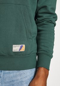 Felpa verde con tasca frontale, polsini e orlo a costine. Presenta un'etichetta colorata con design geometrico e testo sulla tasca.