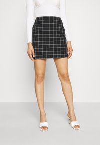 Femme portant une mini-jupe à carreaux noir et blanc, un haut à manches longues blanc et des chaussures ouvertes à talons hauts blanches, se tenant sur un sol gris.