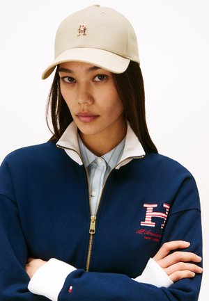 Cappello beige con logo frontale, indossato con una giacca zip blu navy con colletto bianco e dettagli ricamati rossi su una maglietta a righe.