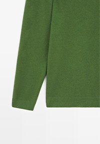Manche et ourlet inférieur d’un pull vert tricoté avec poignets et ceinture côtelés sur fond blanc.