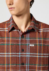 Camicia a quadri marrone con linee multicolori in arancione, blu e verde. Presenta una chiusura a bottoni, colletto e un piccolo etichetta logo Wrangler.