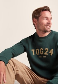 Green sweatshirt featuring gold embroidered text "TOG24 EST. 1958," with ribbed cuffs and neckline, paired with beige trousers.