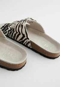 Hvide og sorte zebra-stribede slip-on sandaler med en polstret sål og korkbund. Har en spænde og udskæring på stroppen.