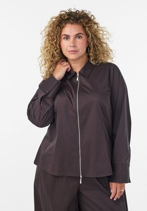 Frau mit lockigem Haar, die ein dunkles langärmeliges Zip-up-Shirt und passende Hose trägt und mit neutralem Gesichtsausdruck nach vorn schaut.