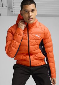Lys orange pufferjakke med kviltet tekstur, sorte sidelapper, frontlynlås og hætte. Har et hvidt Puma-logo på brystet.