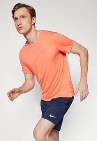 Herre træningsoutfit bestående af en lys orange kortærmet t-shirt med et sølvlogo og marineblå shorts med et hvidt logo. Glat stof, tætsiddende design.