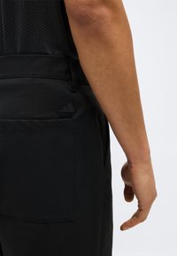 Δεξί χέρι και χέρι ατόμου δίπλα σε τσέπη μαύρου παντελονιού με μικρό μαύρο τριγωνικό λογότυπο σε απλό ανοιχτό φόντο.