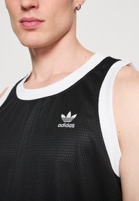 Crni atletski top bez rukava s bijelim rebrastim rubom; sadrži teksturirani uzorak i mali bijeli Adidas logo na prsima.