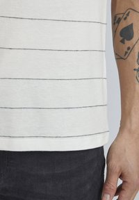 Balta trumpa rankovėmis marškinėliai su horizontaliomis juodomis juostelėmis, su apvaliu kraštu ir minkštu, tekstūruotu audiniu. Matomas rankos tatuiruotė.