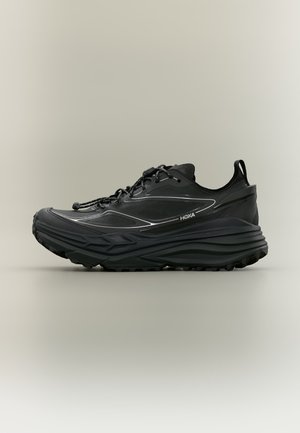 Chaussure de running noire Hoka avec semelle épaisse, languette au talon, détails réfléchissants et fermeture à lacets sur fond neutre.