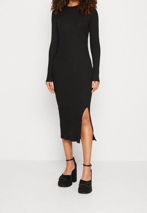 Abito nero a costine a maniche lunghe con spacco laterale fino al ginocchio. Presenta una silhouette aderente e uno scollo tondo. Abbinato a scarpe con platform e cinturino alla caviglia.