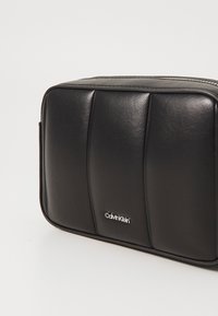 Borsa rettangolare in pelle nera con design trapuntato, dotata di un logo argentato e una texture liscia. Chiusura con zip e lati piatti.