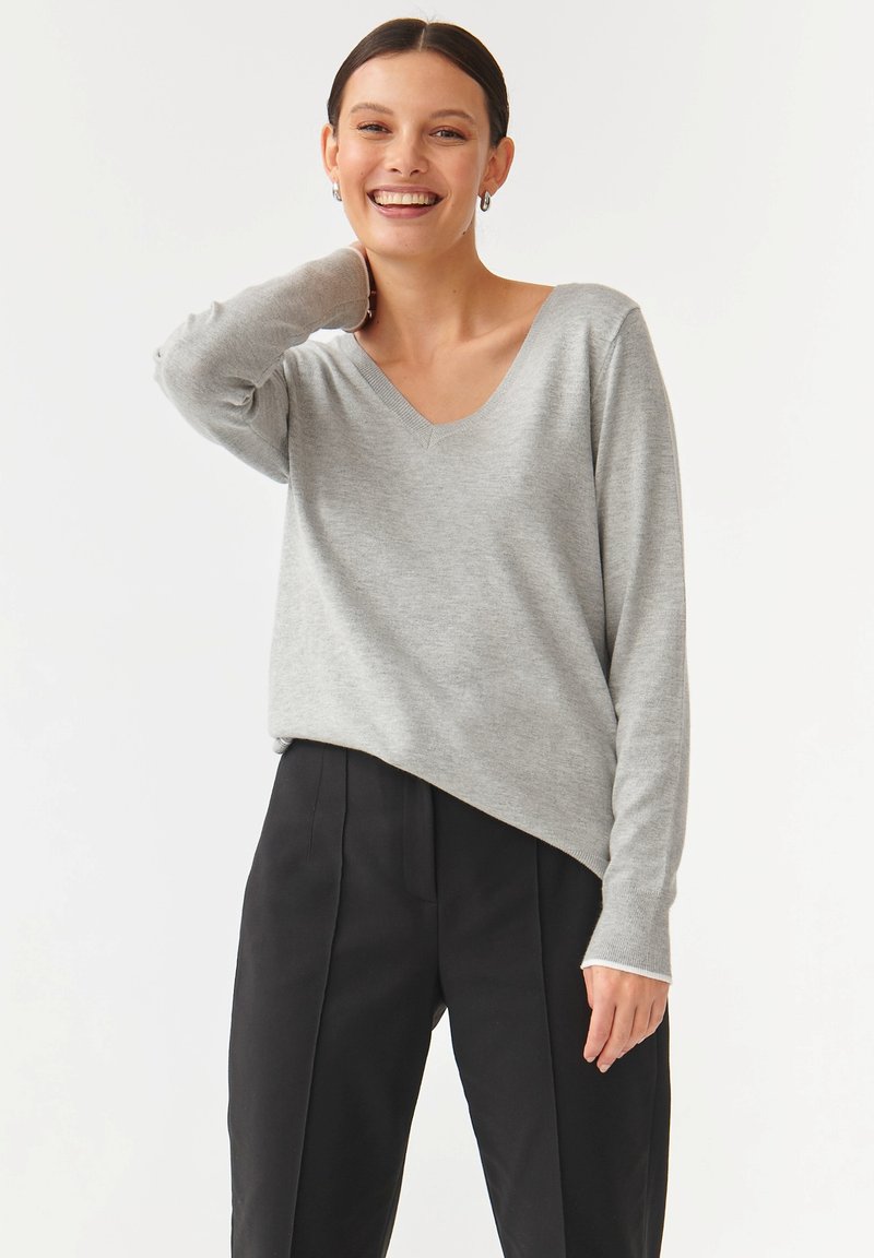 Grå V-hals sweater i blødt strik, med lange ærmer og en let asymmetrisk kant, parret med højtaljede sorte bukser.