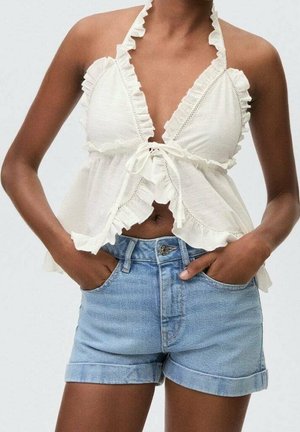 Une femme porte un haut halter blanc à volants noué devant et un short en jean bleu clair taille haute, les mains dans les poches.