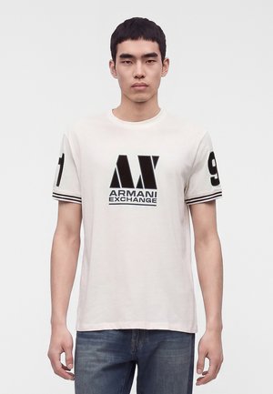 Νέος άνδρας που φορά ένα λευκό T-shirt Armani Exchange με μαύρο λογότυπο και αριθμούς στα μανίκια, συνδυασμένο με μπλε τζιν, στέκεται μπροστά σε λευκό φόντο.