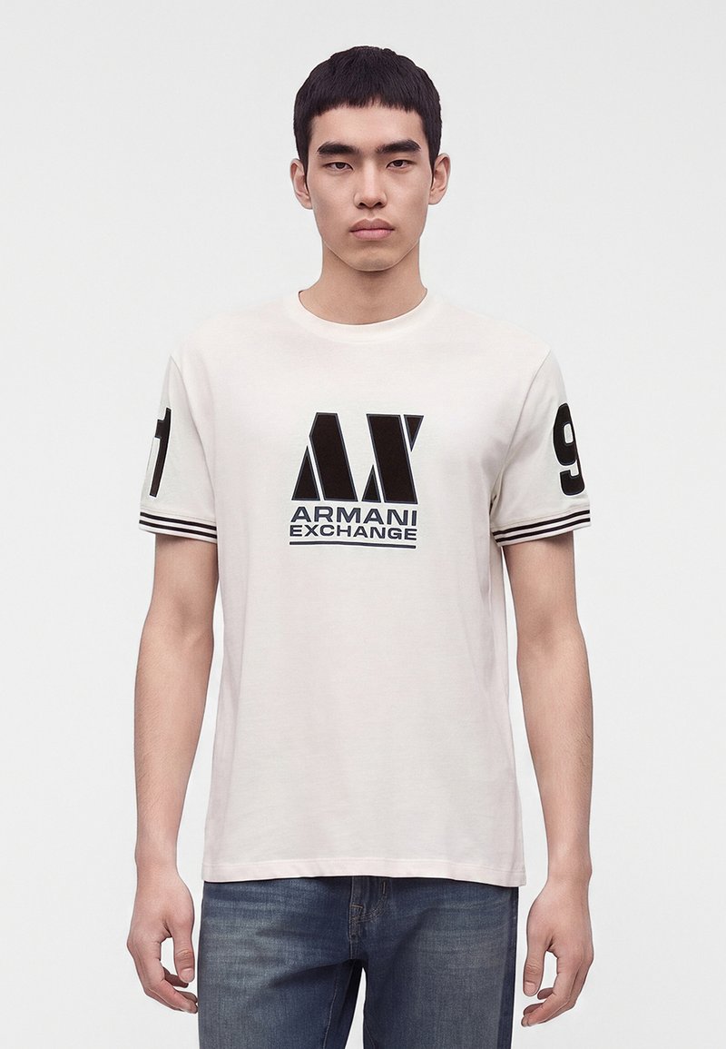 Νέος άνδρας που φορά ένα λευκό T-shirt Armani Exchange με μαύρο λογότυπο και αριθμούς στα μανίκια, συνδυασμένο με μπλε τζιν, στέκεται μπροστά σε λευκό φόντο.