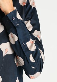 Sleeve of a navy blue shirt with large light beige and peach floral patterns, buttoned cuff visible, hand partially covering face.