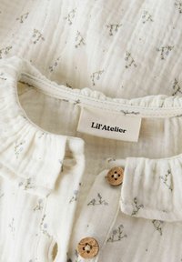 Morbido capo per neonato bianco con delicata stampa floreale viola, bottoni di legno, collo arricciato e etichetta "Lil'Atelier" all'interno della scollatura.