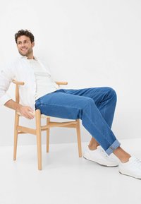 Sedia di legno chiaro con sedile intrecciato, un uomo in camicia bianca, t-shirt bianca, jeans blu e sneakers bianche, seduto in modo informale.