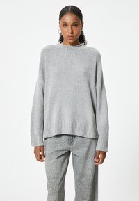 Hellgrauer, gestrickter Pullover mit Rundhalsausschnitt, überschnittenen Schultern und geripptem Saum; kombiniert mit grauen Hosen mit Nietenverzierung.