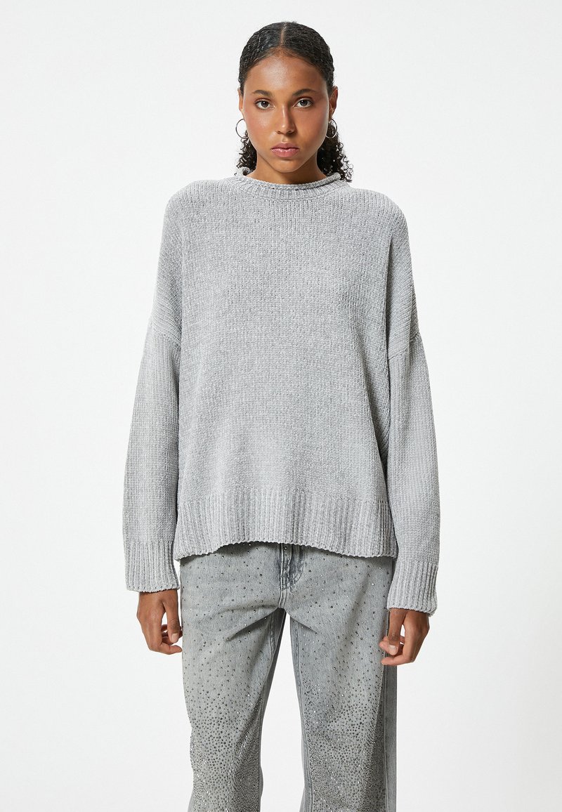 Hellgrauer, gestrickter Pullover mit Rundhalsausschnitt, überschnittenen Schultern und geripptem Saum; kombiniert mit grauen Hosen mit Nietenverzierung.