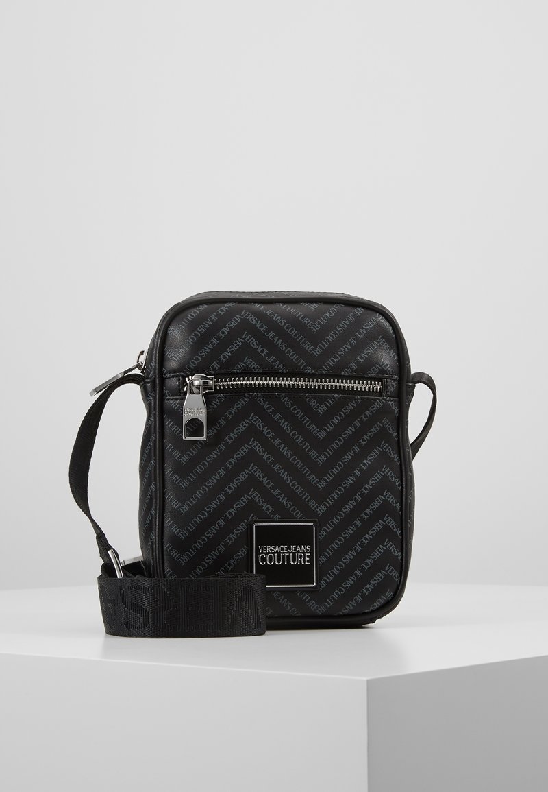 Borsa a tracolla nera con finitura testurizzata, motivo logo ripetuto, chiusura con zip e tracolla regolabile. Presenta dettagli in metallo.