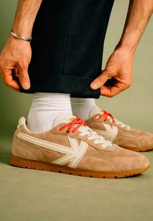 Persoon rolt een zwarte broek op om witte sokken en beige sneakers met witte en rode veters te laten zien op een lichtgroene achtergrond.