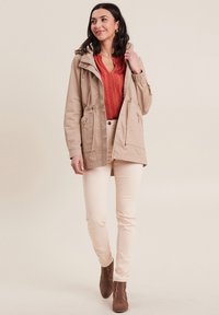 Veste légère beige avec capuche, taille à cordon, et deux poches zippées à l'avant. Portée sur un blouse texturée orange et un pantalon crème. Bottines brunes.