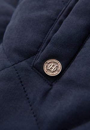 Marinefarbenes Steppgewebe mit sichtbaren Nähten, ausgestattet mit einem runden Bronzeknopf, der mit "Ombre Heritage" geprägt ist. Texturierte Oberflächenstruktur ist zu erkennen.