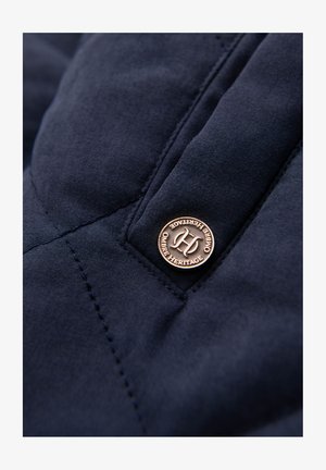 Marinefarbenes Steppgewebe mit sichtbaren Nähten, ausgestattet mit einem runden Bronzeknopf, der mit "Ombre Heritage" geprägt ist. Texturierte Oberflächenstruktur ist zu erkennen.