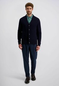 Marinefarbener Strickcardigan mit fünf Knöpfen, gerippten Bündchen und Saum, getragen über einem mintgrünen Polo und dunklen Hosen, kombiniert mit braunen Schuhen.