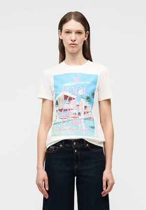 Kobieta w białym t-shircie z pastelową grafiką sceny przy basenie i różowym napisem „Pool & Social Club Versace Jeans Couture”, w połączeniu z ciemnymi dżinsami.