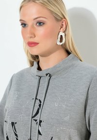 Felpa grigia con scollatura alta, caratterizzata da una grafica con lettering nero e dettagli in strass. Il modello indossa grandi orecchini bianchi astratti.