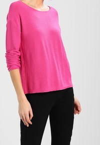 Różowy sweter o luźnym kroju, z dekoltem typu scoop i długimi rękawami. Wykonany z miękkiego, teksturowanego materiału. Połączony z czarnymi, dopasowanymi spodniami.