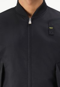 Giubbotto bomber nero realizzato in tessuto liscio, dotato di zip frontale, collo a costine e un'etichetta con testo giallo sul lato sinistro del petto.