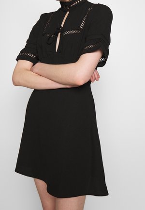 Woman wearing a black short-sleeve dress with lace panels on sleeves and chest, standing with arms crossed against plain background.