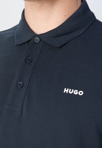 Granatowa koszulka polo wykonana z teksturowanej tkaniny, z trzema guzikami na plastrze i białym logo "HUGO" na lewej piersi.