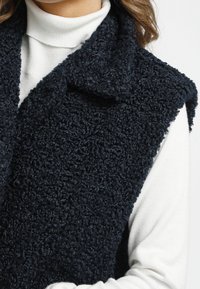 Granatowa, puszysta kamizelka wykonana z teksturowanej tkaniny, z kołnierzem i bez rękawów, nałożona na białą bluzkę z golfem.