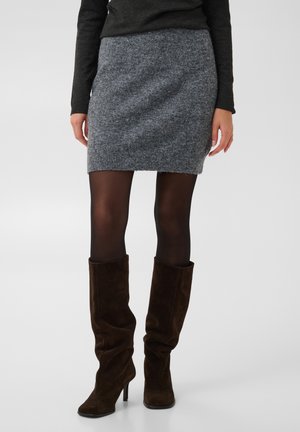 Femme portant une jupe en maille grise, des collants noirs transparents, des bottes marron foncé à talons hauts et amples, et un haut à manches longues foncé, debout sur un fond uni.