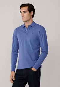 Camisa polo de manga larga en tejido azul claro liso, con un cuello, tapeta de tres botones y un detalle de logo en el pecho.