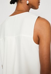 Top senza maniche bianco realizzato in un tessuto leggero e morbido. Dettagli notevoli includono una piega sul retro e un design del colletto pulito.