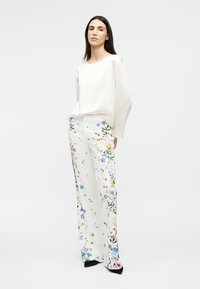 Γυναίκα που φοράει ένα άνετο άσπρο μακρυμάνικο μπλουζάκι και φαρδιά άσπρη παντελόνα με πολύχρωμα λουλουδάτα σχέδια, στέκεται μπροστά σε λευκό φόντο.