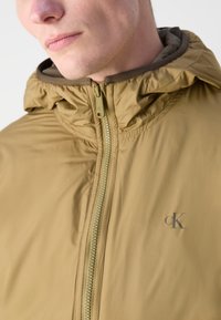 Leichte, strukturierte Jacke in Beige mit Kapuze und einem Reißverschluss vorne. Verfügt über ein dezentes Logo auf der linken Seite und ein kontrastierendes Innenfutter.
