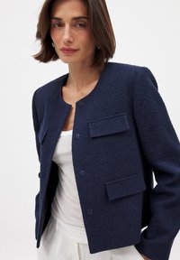 Jūrinės tekstūros švarkas su apvaliu kaklu, priekyje užsegamas mygtukais ir dviem kišenėmis, derinamas su balta palaidine po juo.