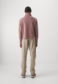 Sudadera de polar en color rosa polvo, con cuello alto y diseño recortado. Combinada con pantalones beige y zapatillas multicolor. Textura suave.
